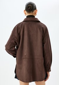 Chaqueta de ante sintético marrón con mangas largas, un cuello y ligeras aberturas laterales. Textura suave, corte ligeramente oversize.