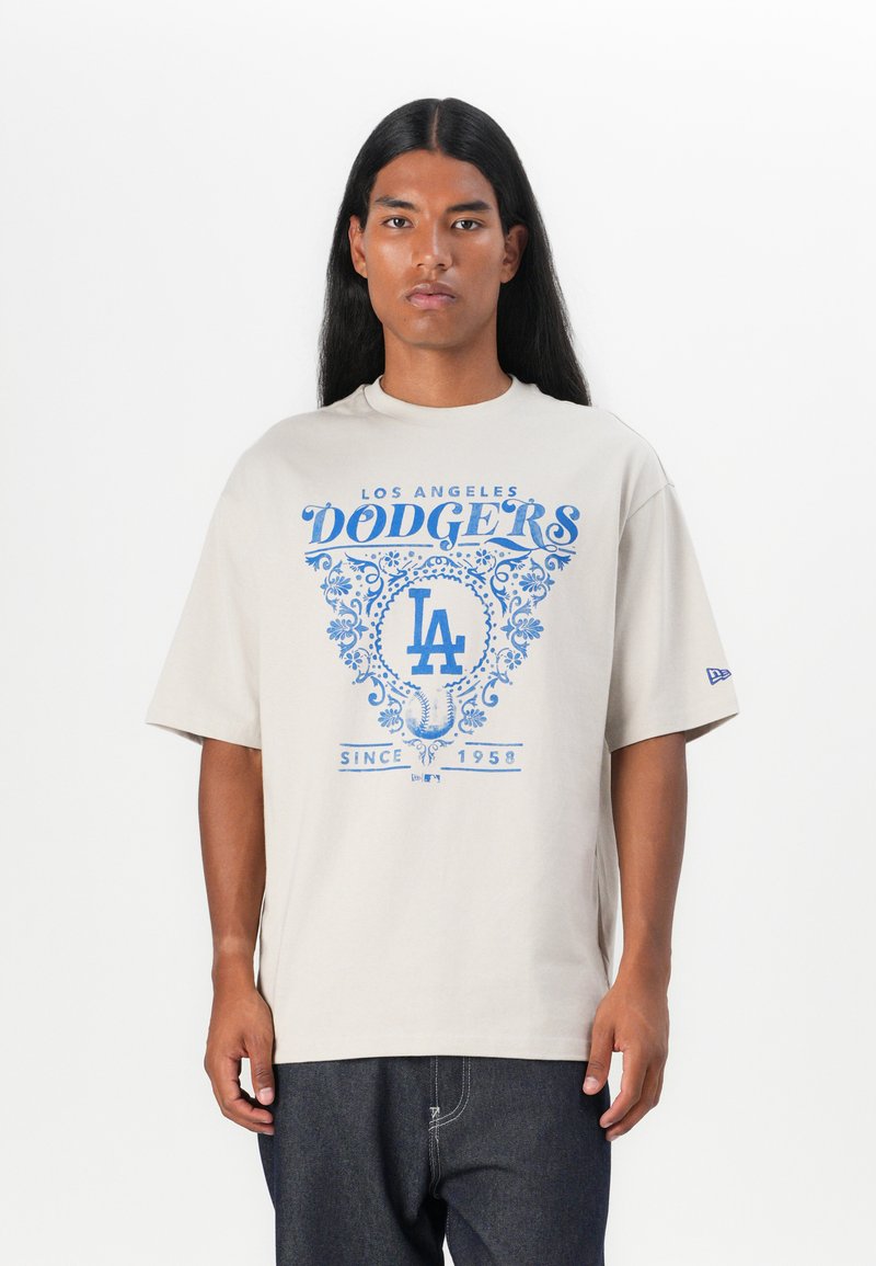 Beige kokvilnas t-krekls ar zilu Los Angeles Dodgers logo, kas satur dekoratīvus elementus, brīvu piegriezumu un īsām piedurknēm.