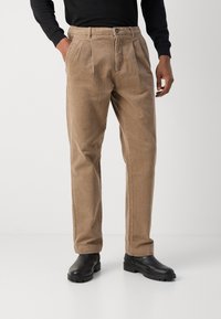 Pantalones de pana beige con un corte relajado, que cuentan con un cierre de botón, pliegues en el frontal y una textura suave, combinados con botas negras.