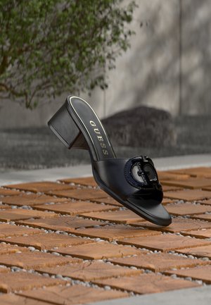 Μαύρο mule Guess με ψηλό τακούνι και ανοιχτή μύτη, με λεπτομέρεια λογότυπου, εμφανίζεται να αιωρείται πάνω από ξύλινα πλακάκια με σχέδιο σε εξωτερικό χώρο με θολό πράσινο φόντο.