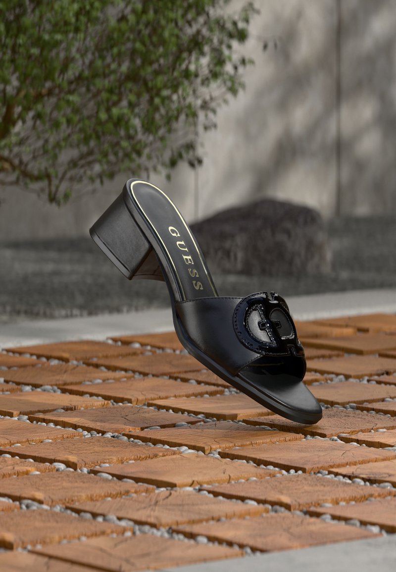 Μαύρο mule Guess με ψηλό τακούνι και ανοιχτή μύτη, με λεπτομέρεια λογότυπου, εμφανίζεται να αιωρείται πάνω από ξύλινα πλακάκια με σχέδιο σε εξωτερικό χώρο με θολό πράσινο φόντο.