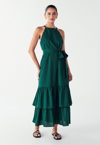 Grünes Leinenkleid mit einem taillierten Oberteil, Krawattenbunddetails und einem gestuften Rüschensaum. Mit Spaghettiträgern und einer lässigen Silhouette.