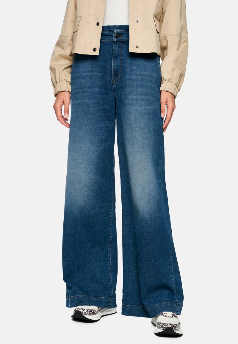 Wide-leg denim jeans in medium blue, featuring a faded wash and a straight cut. Paired with white sneakers and a cropped beige jacket.