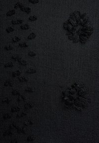 Musta kangas, jossa on pieniä kohollaan olevia pyöreitä ja epäsäännöllisiä nyppyläkuvioita, jotka muodostavat hienovaraisen kukkakuvion.