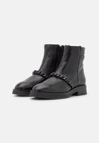Botas de tobillo de cuero negro con cremalleras laterales, suelas de goma planas y cadena metálica negra decorativa en la parte frontal.