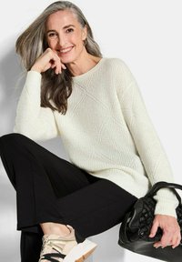 Weißer Strickpullover mit strukturiertem Rautenmuster, schwarze weit geschnittene Hose und beigem Sneaker. Das Outfit umfasst eine gewebte schwarze Handtasche.