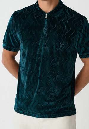 Homme portant un polo texturé vert foncé à manches courtes avec un col à zip quart, associé à un pantalon clair.