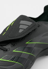 Black soccer shoe with textured upper, green accents, and gray stripes. Features a lace-up closure and a fold-over flap at the top.