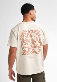 Crèmekleurig katoenen T-shirt met een groot, abstract bruin bloemdesign op de achterkant met tekst onder de afbeelding.