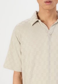 Bež majica polo z teksturiranim kariranim vzorcem, kratkimi rokavi in ovratnikom, z rahlo sijočo površino in ohlapnim krojem.