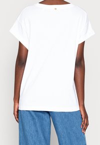 White cotton t-shirt with a round neckline, drop shoulder sleeves, and a small gold accent at the back. Simple, relaxed fit design.