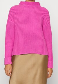 Pull en tricot fuchsia avec col montant, manches larges et texture côtelée. Associé à une jupe en satin doré, présentant une coupe décontractée.