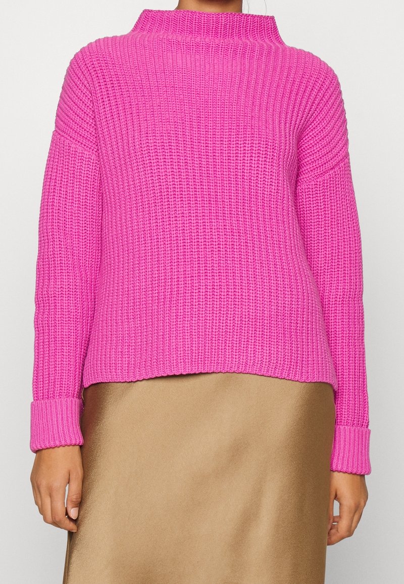 Pull en tricot fuchsia avec col montant, manches larges et texture côtelée. Associé à une jupe en satin doré, présentant une coupe décontractée.