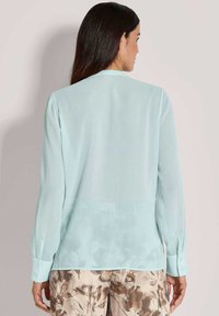 Himmelblaue Bluse mit langen Ärmeln aus einem transparenten Material, mit Knopfmanschetten und lässigem Schnitt. Getragen über floral gemusterten Hosen.