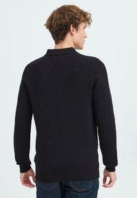 Mies nuori kihara hius, päällään musta pitkähihaiste takki ja siniset farkut, seisoo selin vaaleaa taustaa vasten.