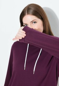Junge Frau mit langen braunen Haaren, die einen lila Kapuzenpullover trägt und den unteren Teil ihres Gesichts mit ihrem Ärmel bedeckt, vor einem weißen Hintergrund.