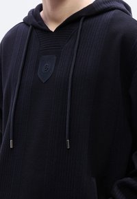 Μαύρο φούτερ με κουκούλα από υφασμάτινη ύφανση, με κάθετες ραβδώσεις, έμβλημα ασπίδας στο στήθος και κορδόνια με μεταλλικές άκρες.