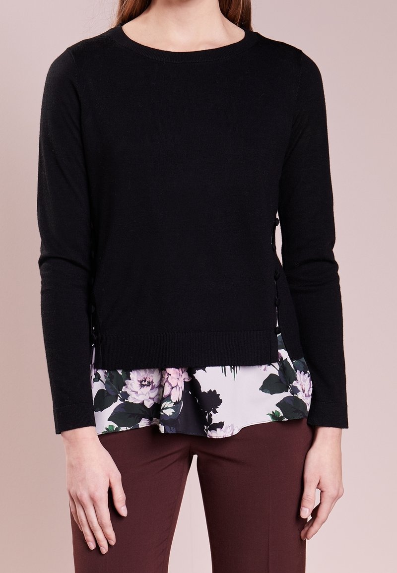 Černý svetr s dlouhými rukávy a kulatým výstřihem, který má boční knoflíky a vrstvený květinový potisk na lemu v odstínech růžové a zelené.