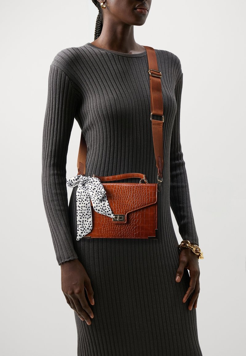 Femme portant une robe côtelée gris foncé et un sac bandoulière marron texturé avec une écharpe blanche à motifs attachée à la poignée.