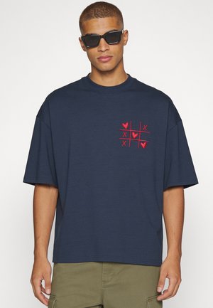 Homme portant des lunettes de soleil noires et un t-shirt bleu marine avec un motif rouge de morpion avec des cœurs et des X sur la poitrine.