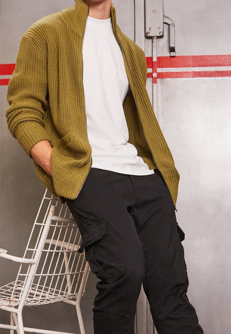 Olivově zelený žebrovaný cardigan na zip přes bílý tričko, doplněný černými cargo kalhotami. Oba kousky mají volný střih a jemné textury.