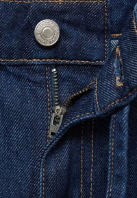 Donkerblauwe denimstof met zichtbare stiksels, een metalen knoop met het label "Original Penning Collection" en een gedeeltelijk open zwarte rits.