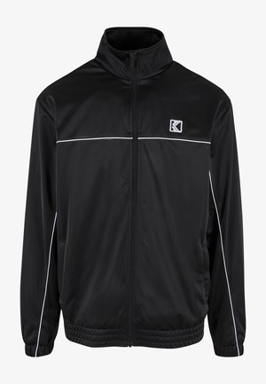 Schwarze Sportjacke aus glattem, leichtem Stoff. Mit hohem Kragen, Reißverschluss vorne, weißen Akzentpaspeln und seitlichen Taschen.