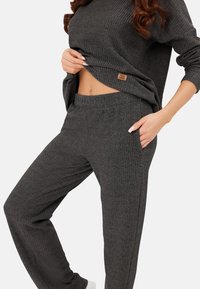 Kobieta ubrana w ciemnoszary, prążkowany sweter i zestaw spodni, podnosząca lekko sweter, aby pokazać brzuch, z ręką w kieszeni spodni.
