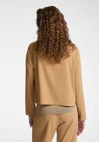 Beige měkká látková bunda s volným střihem, dlouhými rukávy a žebrovaným lemem, zobrazená zezadu s viditelnými kudrnatými vlasy.