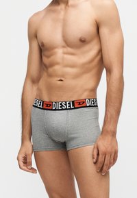 Torse masculin athlétique portant un boxer Diesel gris avec ceinture noire et logo rouge sur un fond clair uni.
