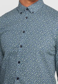 Camisa azul de botones con un pequeño patrón abstracto de puntos blancos y verde claro, llevada por una persona mostrada desde el cuello hasta la mitad del torso.