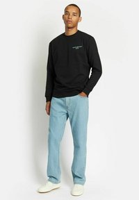 Svart genser med rund hals og turkise bokstaver, sammen med lyseblå denimjeans og hvite joggesko, stående mot en ensfarget bakgrunn.