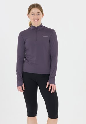 Pitkähihainen, vetoketjullinen urheilu toppi tumman violetista kankaasta, yhdistetty mustiin capri-leggingseihin. Sisältää tekstuuripinnan ja logoyksityiskohdan.