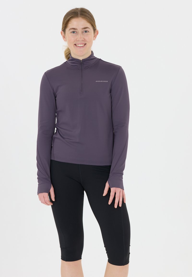 Pitkähihainen, vetoketjullinen urheilu toppi tumman violetista kankaasta, yhdistetty mustiin capri-leggingseihin. Sisältää tekstuuripinnan ja logoyksityiskohdan.