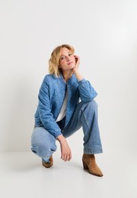 Chaqueta bomber acolchada azul claro con puños de canalé, combinada con jeans azul claro y botas de ante marrón hasta el tobillo. Diseño minimalista, brillo sutil.