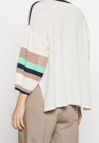 Krämfärgad topp med trekvarts ribbade ärmar som har horisontella ränder i beige, grön och blå, med en avslappnad passform och mjuk textur.