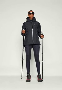 Veste imperméable noire avec fermeture éclair, associée à des leggings foncés et des chaussures de randonnée avec des accents rouges. Bâtons de randonnée dans les deux mains.
