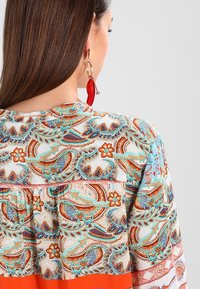 Patterned blouse with paisley design in turquoise, orange, and beige. Features a gathered hem and contrasting solid orange section at the bottom.