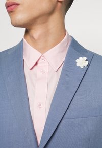 Blazer azul claro com um tecido texturizado, combinado com uma camisa de colarinho rosa pálido com dois botões. Acento de flor branca no lapel do blazer.