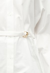 Λευκή υφασμάτινη ζώνη με χρυσή αγκράφα που σφίγγει ένα λευκό πουκάμισο με κουμπιά στη μέση, αναδεικνύοντας τις λεπτομέρειες και την υφή του ρούχου.