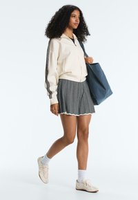 Mladá žena kráča, oblečená v krémovej bunde na zips, sivej skladanej sukni, bielych ponožkách a teniskách, s veľkou modrou taškou cez rameno.