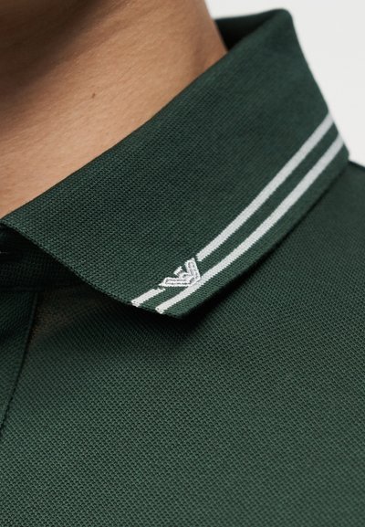 Col polo vert foncé avec deux bandes blanches et un petit logo blanc brodé près du bord, porté sur le cou d'une personne.