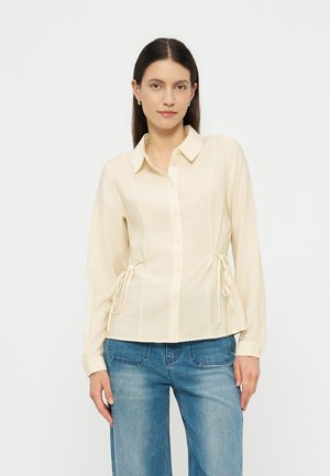 Γυναίκα με μακριά σκούρα μαλλιά φοράει κρεμ πουκάμισο με κουμπιά και δεσίματα στο πλάι και μπλε τζιν, στέκεται μπροστά σε απλό λευκό φόντο.