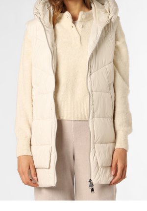 Femme portant un gilet matelassé à capuche écru sur un pull en tricot crème et un pantalon beige clair, les mains détendues le long du corps, fermeture éclair partiellement ouverte.