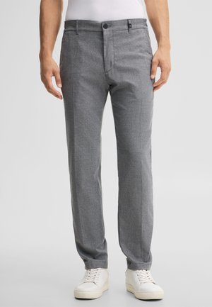 Mężczyzna ubrany w szare spodnie typu slim fit i białe trampki stoi na jednolitym tle.