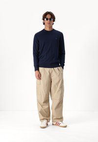 Námornícký svetr s dlouhým rukávem, béžové cargo kalhoty, bílé tenisky s červenými pruhy. Model v brýlích stojí na bílém pozadí.