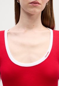 Gros plan d'une personne portant un haut rouge à encolure dégagée avec un bord blanc et un petit logo Calvin Klein près du col.