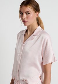 Camicia rosa in satin a maniche corte con colletto, caratterizzata da un delicato motivo a pois e fessure laterali per facilitare il movimento.