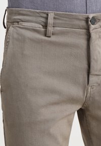 Närbild av beiga byxors midja och övre lårområde som bärs över en blå skjorta, vilket visar knapp, bältesöglor och fickdetaljer.