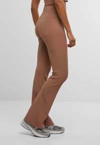 Bruine flared leggings van rekbaar materiaal. Hoog taille ontwerp met een gladde textuur en minimalistische stiksels. Gepaard met grijze sneakers.